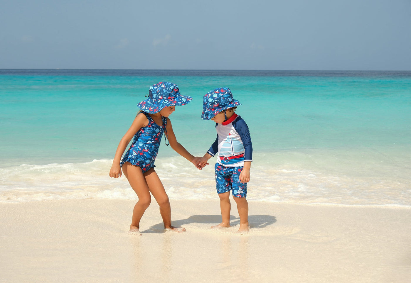 Little boy sitting by the pool wearing a white with whales print flap hat with matching white sleeve with navy blue body and whales print rash guard and swim trunks.