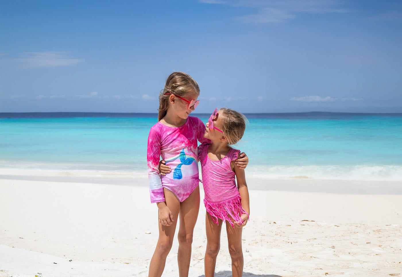 little girl holding a bucket and holding older girl hand as they walk on the beach wearing flap happy swim hats and swimwear in Fantasea Mermaid prints