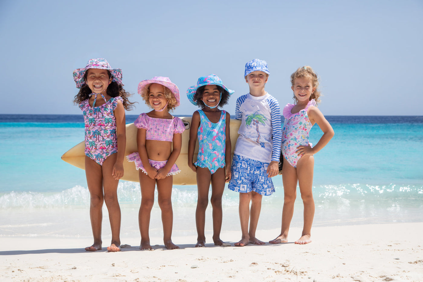 Five young children standing on a beach, smiling and dressed in colorful swimwear and sun hats. The bright blue ocean and clear sky provide a vibrant tropical background, evoking a fun, sunny day by the sea