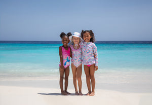 Three young girls standing arm-in-arm on a beach, smiling in colorful swimwear and a sun hat. They are dressed in bright, playful patterns with a vibrant blue ocean and clear sky behind them, capturing a joyful beach moment